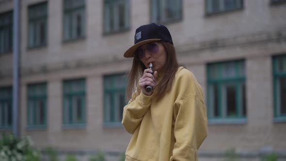 Pretty Young Hipster Woman in Black Cap Yellow Hoodie Vape Ecig. Girl Vape Popular Ecig Gadget