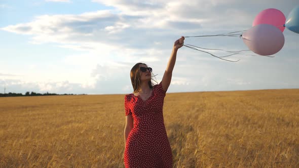 Happy Young Girl with Brown Hair Walking Through Golden Wheat Field and Holding Balloons in Hand