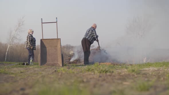 Wide Shot of Mature Caucasian Man Throwing Fallen Leaves Into Fire As His Senior Wife Standing Aside