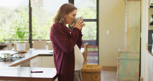 Caucasian pregnant woman standing in kitchen, touching belly and drinking tea