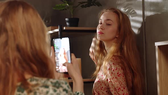 Young Redhead Girl Takes Pictures of His Sister or Girlfriend on a Smartphone That Poses at Home in