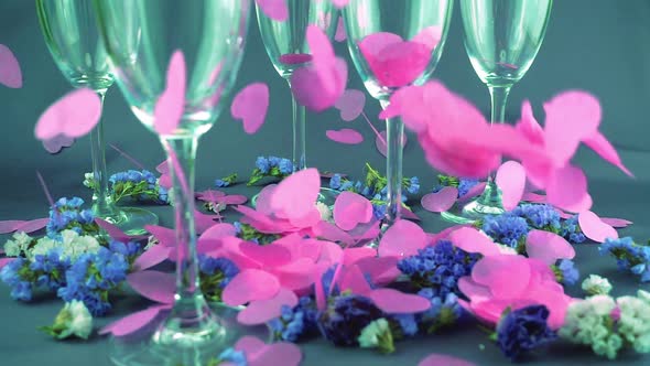 Falling heart shaped confetti on glasses wine and flowers on a purple background. Slow motion.