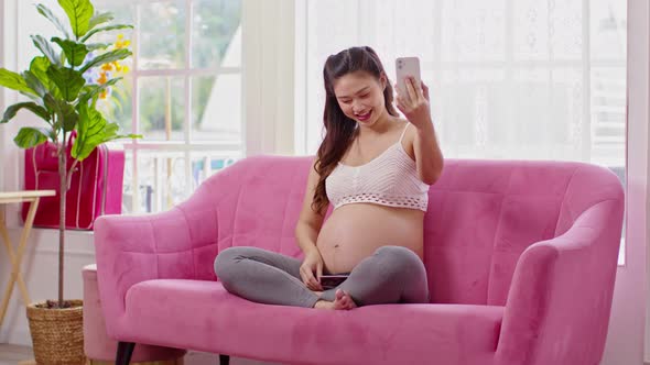 Happy Pregnant Woman sitting on pink couch video call conference on mobile phone with family