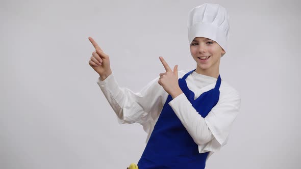 Cheerful Happy Boy in Cook Hat and Apron Pointing Aside Looking at Camera with Toothy Smile