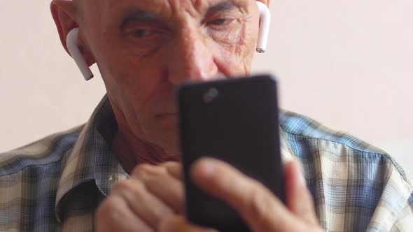 Portrait of a Caucasian pensioner 70-79 in airpods actively uses a smartphone. An elderly serious ma