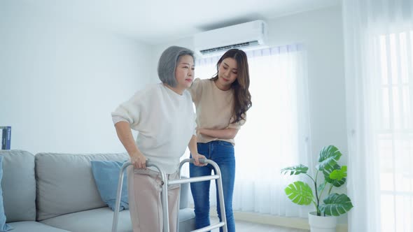 Asian young daughter support old disabled woman doing physical therapy walk with walker at home.