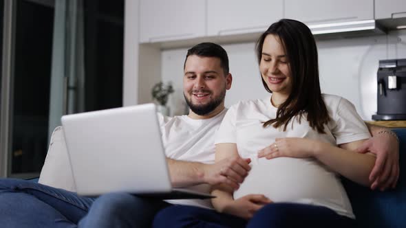 Lovely Pregnant Woman and Man Surf the Net Sitting on the Couch with Laptop