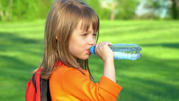 Little Child with School Backpack Drinks Water From Plastic Bottle on Green Lawn in Summer Park