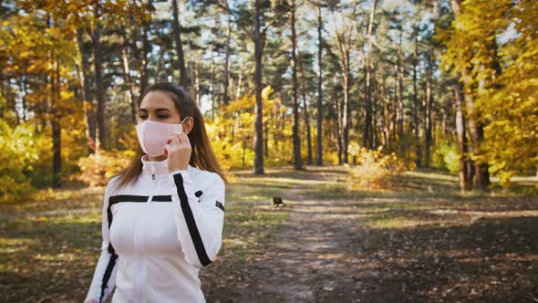 Young Woman in Sportswear is Jogging By Path in Autumn Wood Stopping Taking Off Protective Mask and