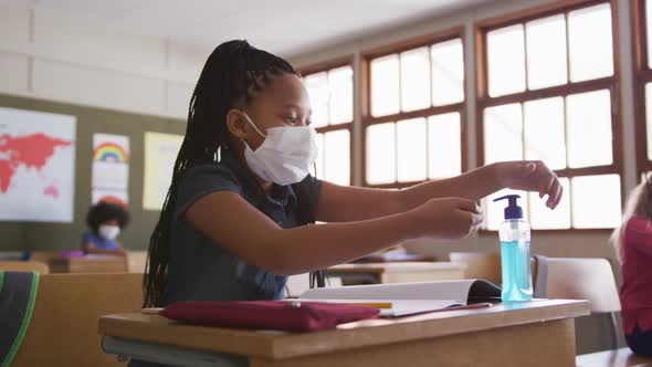 Girl wearing face mask sanitizing her hands while sitting on her desk at school