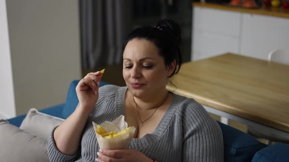Plus Size Woman Eats French Fries with Great Pleasure Sitting on a Couch