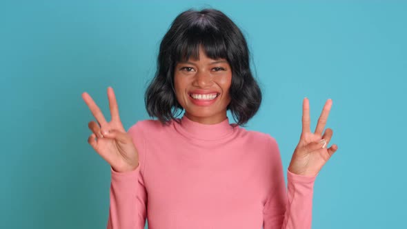 Joyful Dark Skinned Woman Makes Peace Gesture Against Blue Background