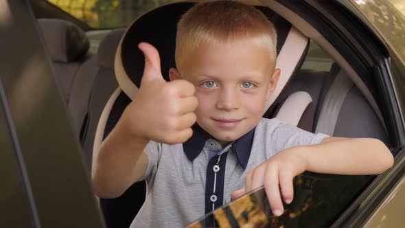 A Kind Cheerful Little Boy Sits in a Car Seat in the Car and Gives a Thumbs Up