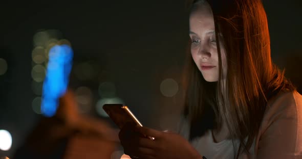 A Young Girl on the Background of the Night City Looks Into the Smartphone Screen