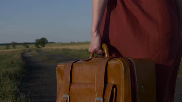 Girl in red dress with suitcase on country road in sunset.