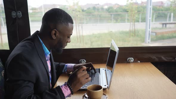 Black Businessman is Working Typing a Message on Laptop Sitting in Summer Cafe