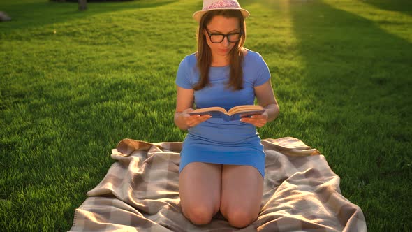 Girl in Glasses Reading Book Sitting on a Blanket in the Park at Sunset