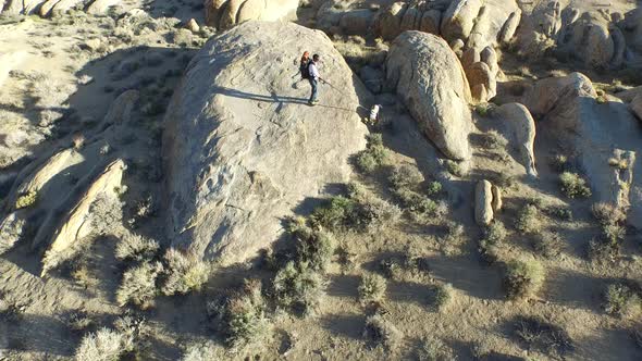 Aerial shot of a young man backpacker standing on a boulder with his dog in a desert mountain range.