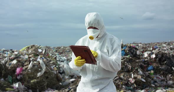Virologist in Protective Suit and Gas Mask Noting in His Tablet at Dump Landfill