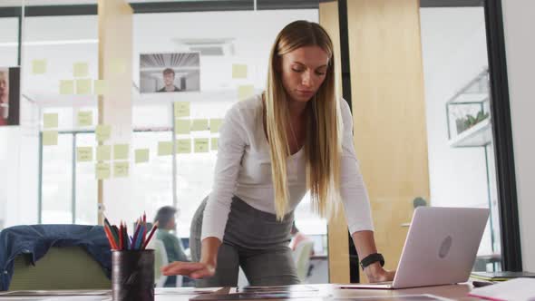 Portrait of caucasian businesswoman standing leaning on desk using laptop and smiling in office