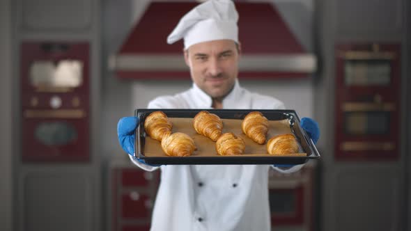 Handsome Baker in Uniform Holding Tray Full of Freshly Baked Croissants at Modern Kitchen