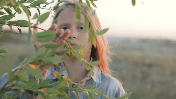 A Little Girl with a Sad Face Looks through the Branches of a Leafy Tree