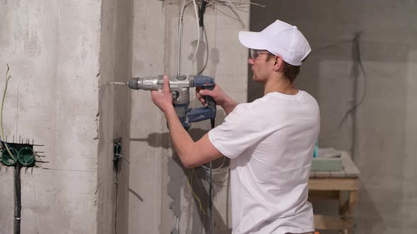 A young worker in a white uniform drills a wall with a punch in apartment