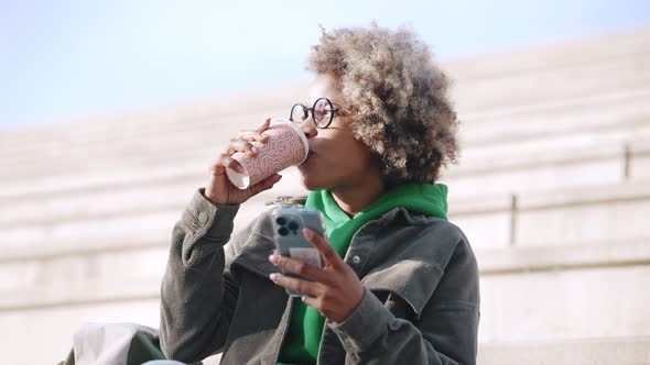 Positive adult African woman wearing casual clothes looking at phone and drinking coffee