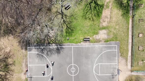 An aerial view over people playing basketball on a court surrounded by dry trees on a sunny day. The