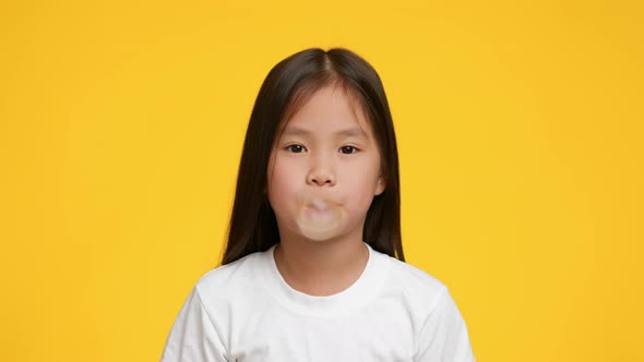 Little Korean Girl Chewing And Blowing BubbleGum Over Yellow Background