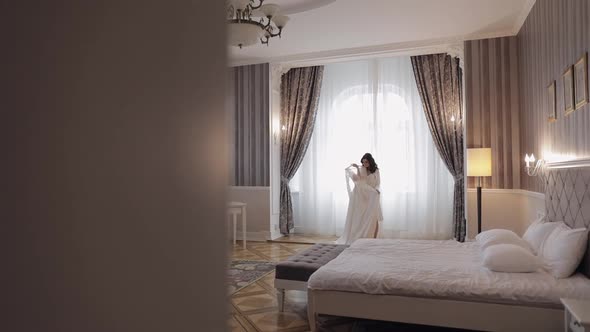 Bride in Lingerie Standing with Her Wedding Dress at Home Near Window White Boudoir Dress and Veil