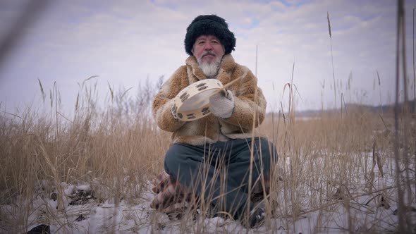 Front View Mongol Shaman Playing Tambourine in Slow Motion Sitting on Snowy Winter Meadow Outdoors