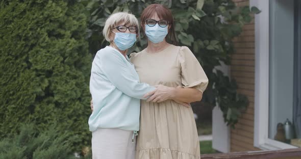 Positive Senior Women in Eyeglasses and Coronavirus Face Masks Hugging and Looking at Camera Smiling