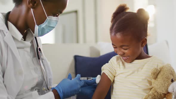 African american female doctor wearing face mask giving covid 19 vaccination to smiling girl at home