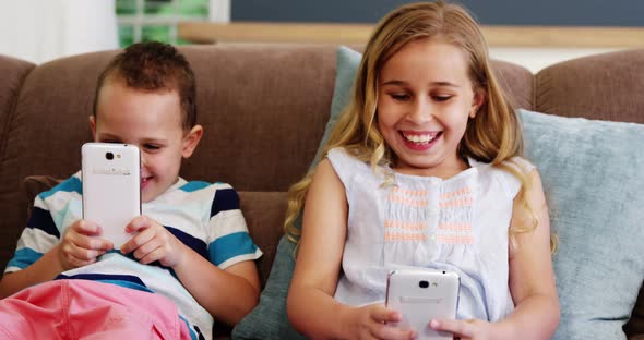 Smiling boy and girl sitting on sofa and using mobile phone