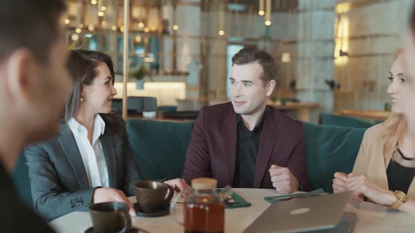 Group of Suit Wearing Colleagues Discussing Working Situation Sitting at Table in Cafe with Laptop