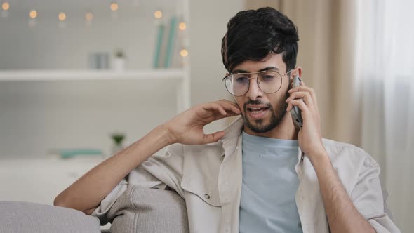 Young Arab Bearded Man Millennial Guy with Glasses Sits on Home Couch Talking on Mobile Phone