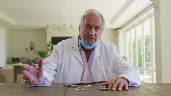 Caucasian senior male doctor wearing lab coat sitting at table talking, giving video consultation