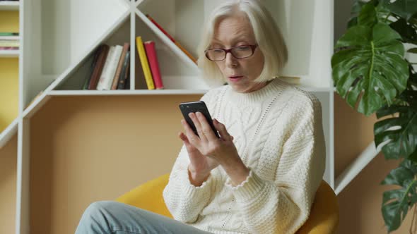 Elderly Woman Is Sitting on the Sofa, Holding a Smartphone, Feeling Very Happy After Receiving an