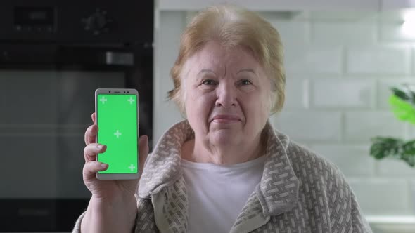 Smiling Elderly Woman Shows Mobile Phone with Green Screen Sitting in Kitchen at Home