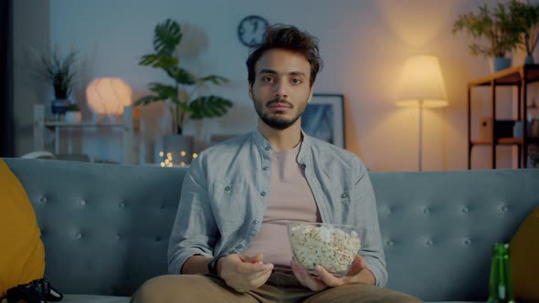Portrait of Pensive Middle Eastern Man Watching TV Eating Popcorn and Thinking in Dark Apartment