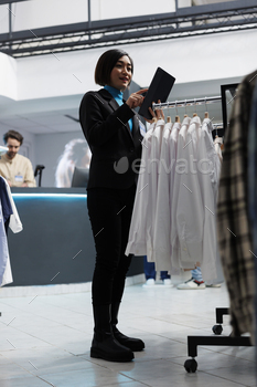 ion inventory using digital tablet. Young asian woman checking apparel stock showcasing on display rack while working in clothing store