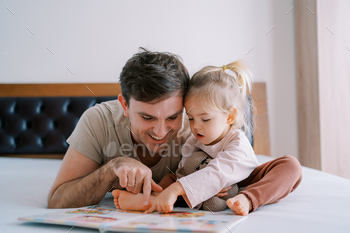 Dad and little girl are reading a book of fairy tales on the bed, running their fingers through the