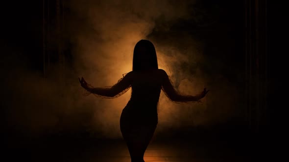 Dark Silhouette of Young Woman Dancing in Dark Smoky Studio Against Background of Yellow Lights