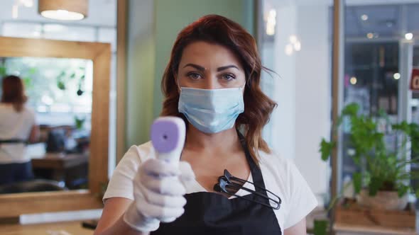 Female hairdresser wearing face mask holding temperature gun at hair salon