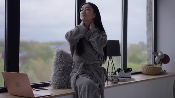 Young Asian Woman Stretching Neck Sitting on Windowsill in Home Office