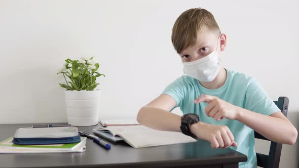 A Young Boy in a Face Mask Looks Up From a School Notebook To the Camera and Taps at His Watch