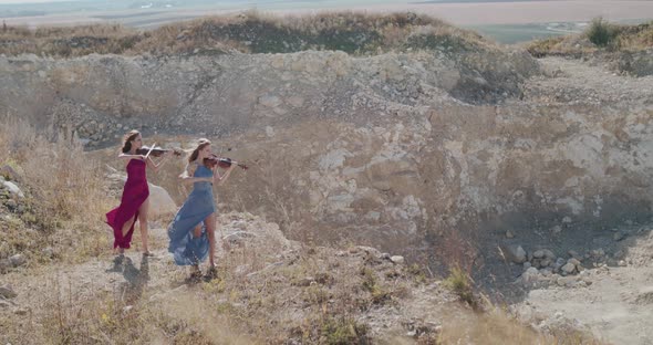 Far View of Two Beautiful Women in Dresses Play Violin Among Cliff in Summer