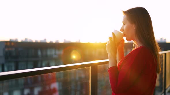 Woman in Red Dress with a Cup of Coffee Standing on the Balcony and Admire the Sunset