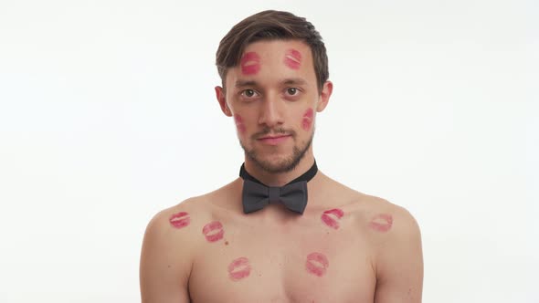 One young brunette man with beard, red kiss imprints or marks on body makes a heart gesture